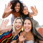 smiling young diverse ladies showing hi sign while taking selfie on terrace