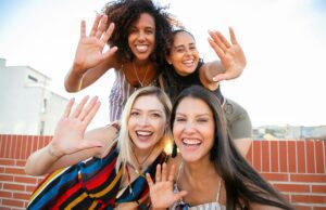 smiling young diverse ladies showing hi sign while taking selfie on terrace